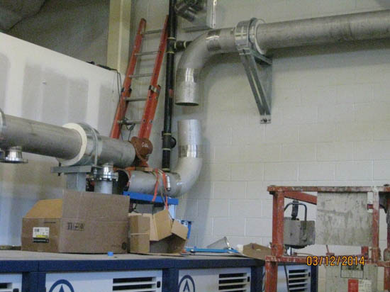 Industrial room with metal pipes, ladders, cardboard boxes, and equipment against a white brick wall.