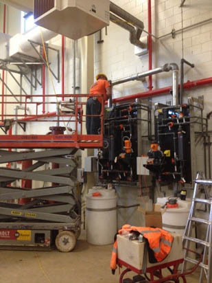 A worker in an orange vest operates equipment on a lift in an industrial setting with tanks and pipes.