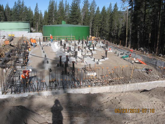Construction site with foundation work, rebar, and workers. Forest and large green tanks in the background.
