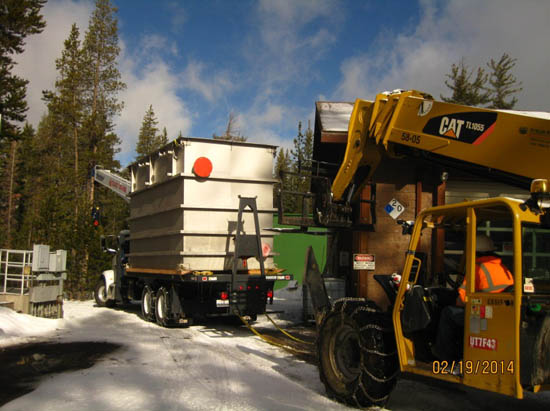 A forklift unloading a large container from a truck in a snowy area with trees in the background.