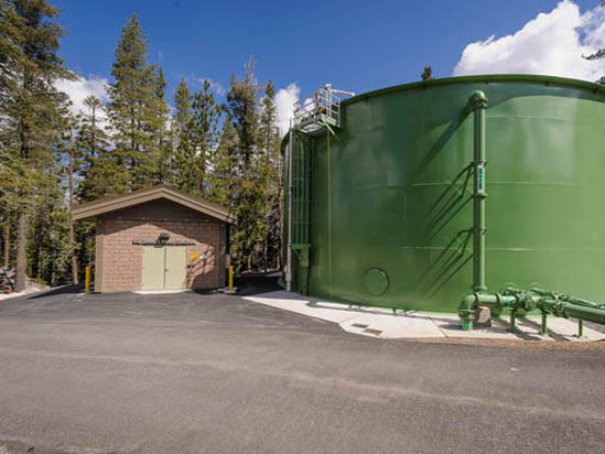 A small building and a large green tank in a forest setting under a blue sky.