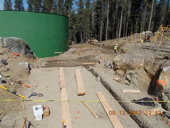 Construction site with wooden beams, a large green tank, and trees in the background.