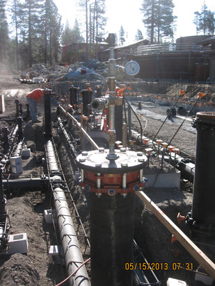 Construction site with pipes and valves, surrounded by dirt and trees.