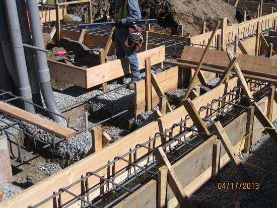 Construction site with wooden forms, rebar, and gravel for a concrete foundation. Workers are present.