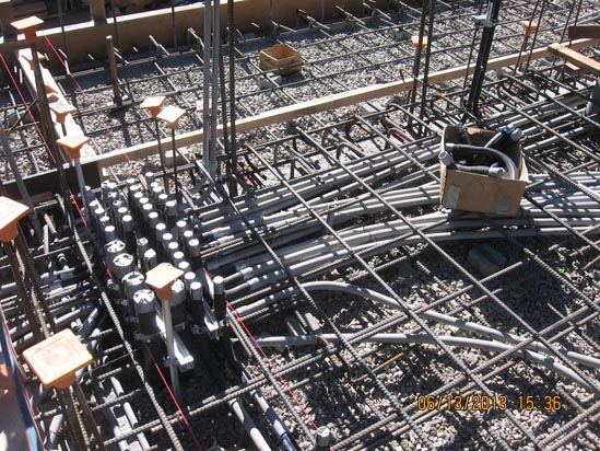 Construction site with metal rebar grid, electrical conduits, and rocks, likely for a concrete foundation.