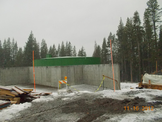 A snowy construction site with concrete walls, safety barriers, a green structure, and piles of materials, surrounded by trees.