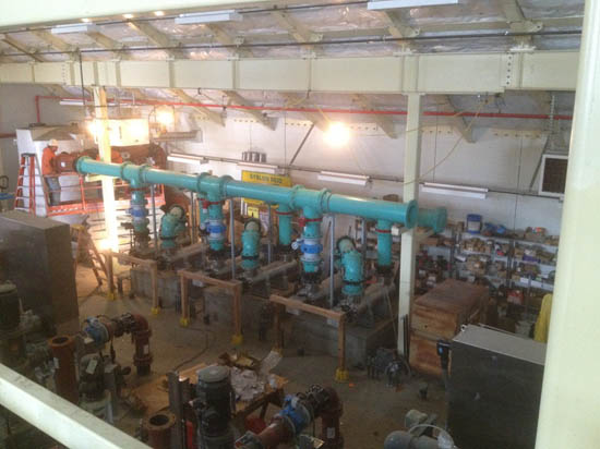 A room with industrial equipment, including large blue and teal pipes, valves, and machinery arranged on a concrete platform.