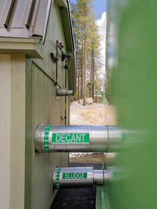 Pipes labeled "DECANT" and "SLUDGE" on the side of a building in a wooded area.