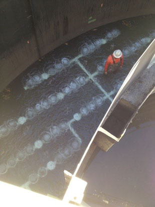 A person in protective gear is standing in a large water-filled industrial tank with visible grid patterns on the surface.