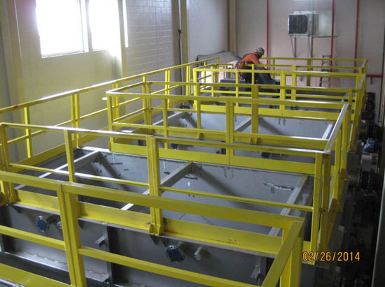 A worker in safety gear stands on large industrial tanks with yellow safety railings.