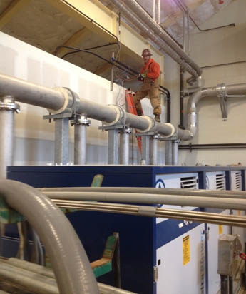 A person in work attire stands on a ladder next to large pipes and industrial equipment indoors.