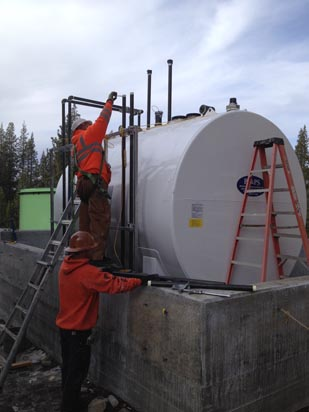Two workers with safety gear are installing or servicing a large white industrial tank on a concrete platform.