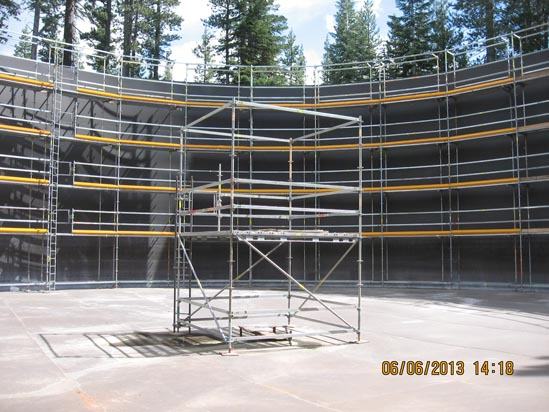 An empty, circular construction site with scaffolding inside and around the perimeter, surrounded by trees.