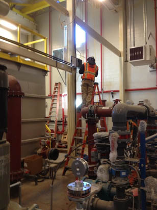 A worker in a safety vest stands on a ladder surrounded by industrial equipment and pipes.