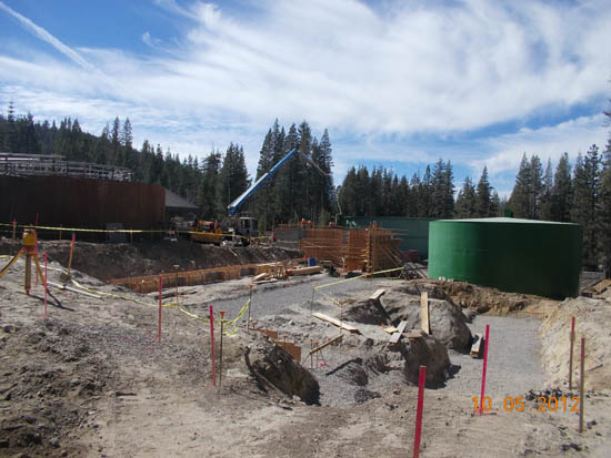 Construction site with large green tank, building materials, and machinery surrounded by trees under a blue sky.