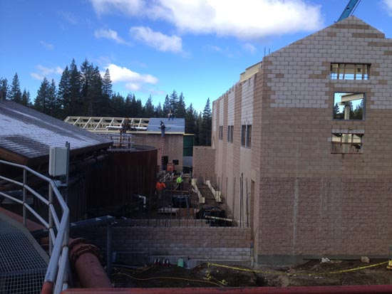 Construction site with unfinished buildings, scaffolding, workers, and equipment surrounded by trees under a blue sky.