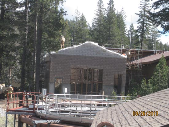 A building under construction surrounded by trees, scaffolding, and a worker on the roof.