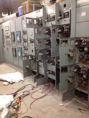 A dismantled electrical control panel with open compartments and exposed wires in an industrial setting.