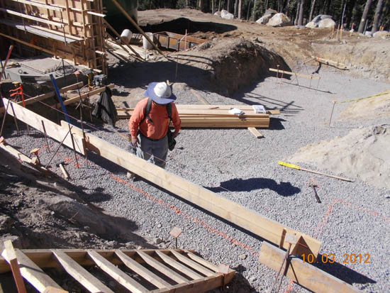 A construction worker at a building site with wooden frames, gravel, and equipment.