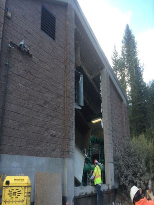 A partially collapsed building with exposed interior, construction workers in helmets and vests around it.
