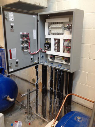 An open electrical control panel installed on a wall with visible wiring and equipment in a mechanical room setting.
