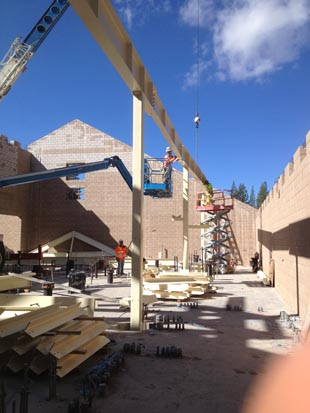 Construction site with cranes lifting beams, surrounded by partially built walls, under a clear blue sky.