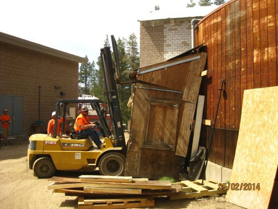 A forklift pushing against a wooden structure, with workers in orange safety vests nearby.