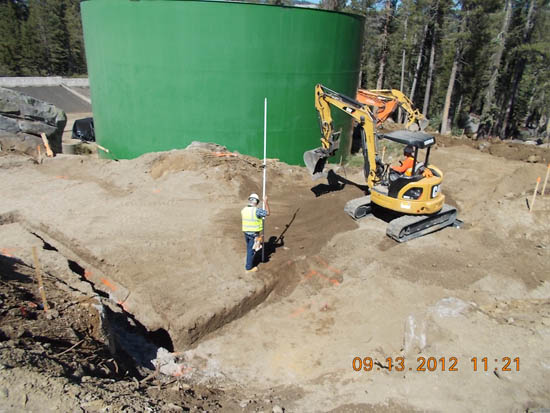 Construction site with a worker holding a pole and an excavator near a large green tank.