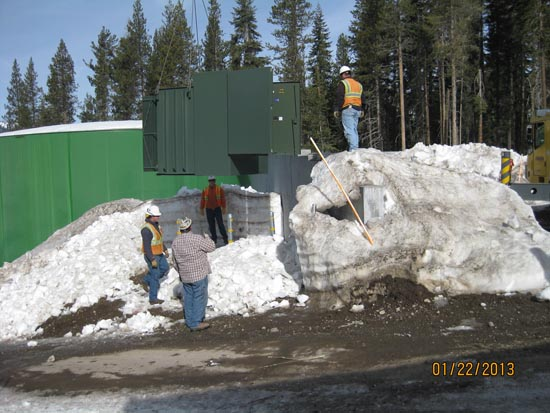 Construction workers in snow working with large green equipment and tools, surrounded by snowy landscape and trees.