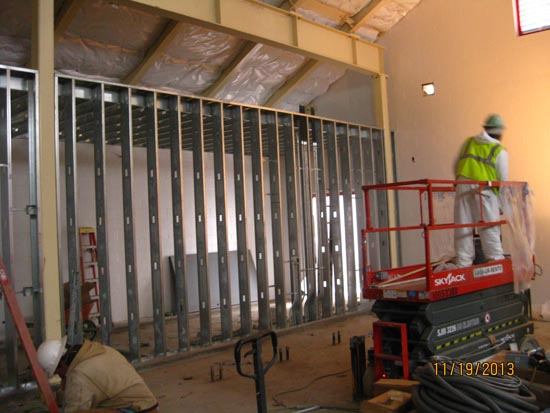 Construction site with workers installing metal studs and insulation. One worker stands on a lift, another is on the ground.