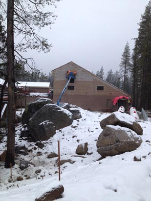 A snow-covered landscape with a building under construction. Two people are on a lift working on the building's roof. Large rocks in the foreground.