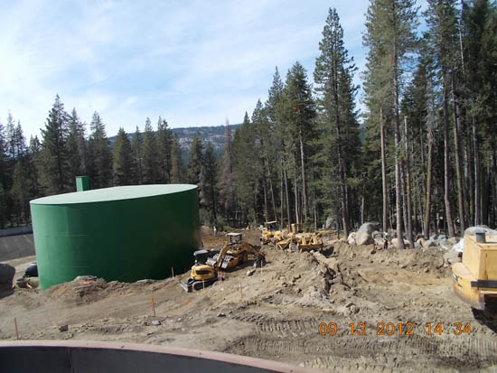Construction site with heavy machinery and a large green storage tank surrounded by trees.