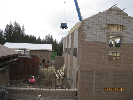 A construction site with a crane lifting equipment, unfinished brick building, and trees in the background.