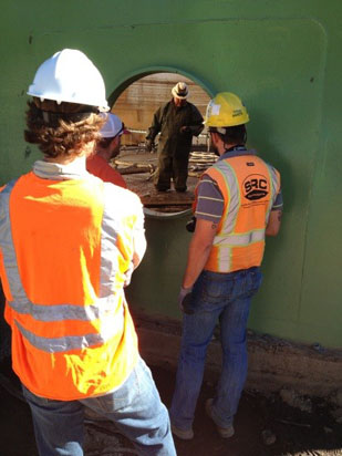 Several construction workers in hard hats and vests observe a person through a circular opening in a large green wall.