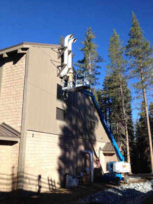 A construction lift reaches up to a building with workers near the roof. Tall trees are in the background under a clear blue sky.