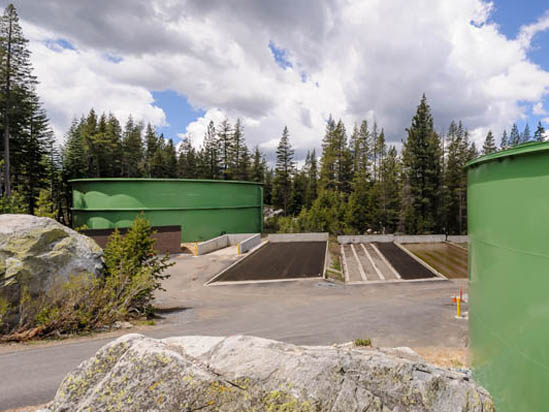 Green storage tanks and concrete structures surrounded by trees and rocky terrain under a cloudy sky.