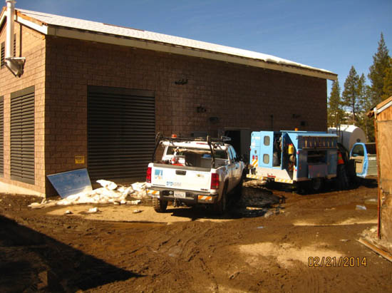 A truck and equipment outside a brick building on a sunny day, with snow and mud on the ground.