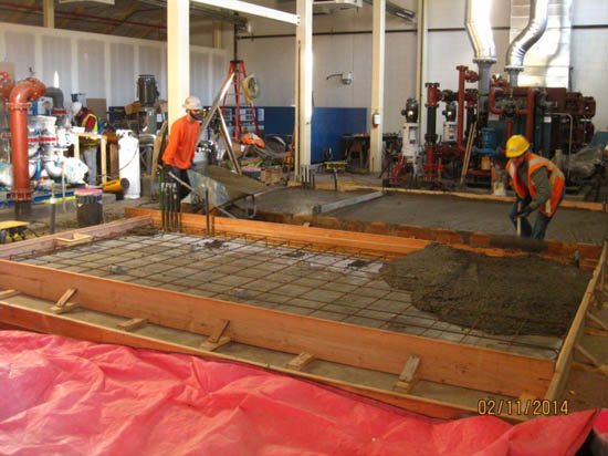 Two construction workers pouring concrete into a rectangular frame with metal reinforcements in an industrial setting.