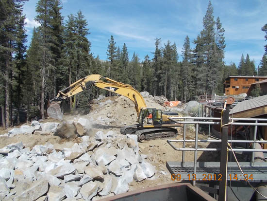 Excavator moving rocks in a forested area with buildings nearby.