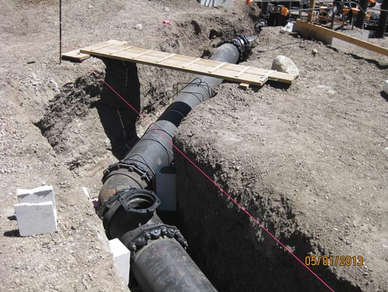 A construction site with large black pipes in a trench, a wooden plank bridge, and ground marked by a red string line.