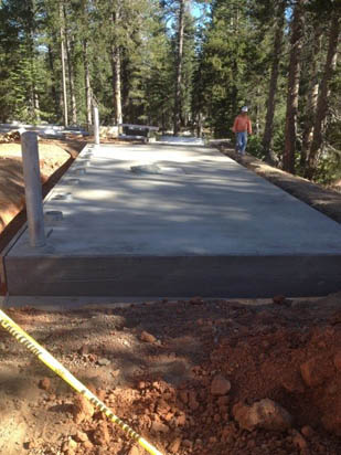 New concrete slab foundation in a forested area with construction tape and a worker nearby.