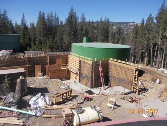 Construction site with scaffolding, ladders, and a large green cylindrical tank, surrounded by trees.
