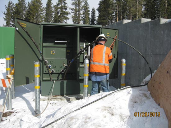 A worker in a hard hat and orange vest operates on electrical equipment outdoors in a snowy area.