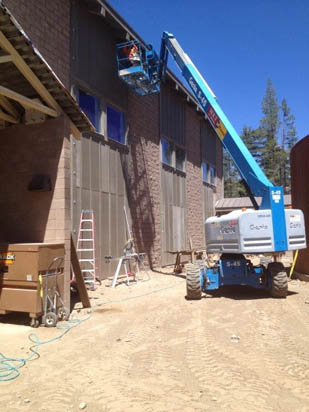 A boom lift is raised against a building under construction, with a person working. Ladders, construction equipment, and trees are nearby.
