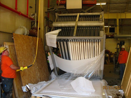 A group of workers manage a large, tilted, industrial machine wrapped in plastic, with wooden boards nearby.