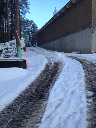 Snowy path with tire tracks beside a concrete building in a wooded area.