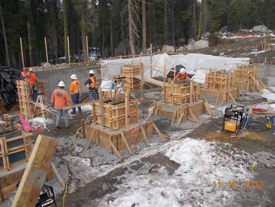 Construction site in a snowy area with workers in safety gear building structures, surrounded by trees.