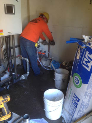 A worker in an orange shirt and hard hat is fixing pipes in a utility room next to buckets and construction materials.