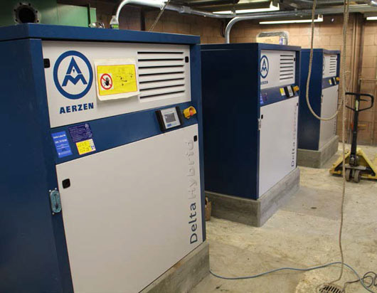Three blue and white industrial machines labeled "Aerzen Delta Hybrid" are lined up in a room.
