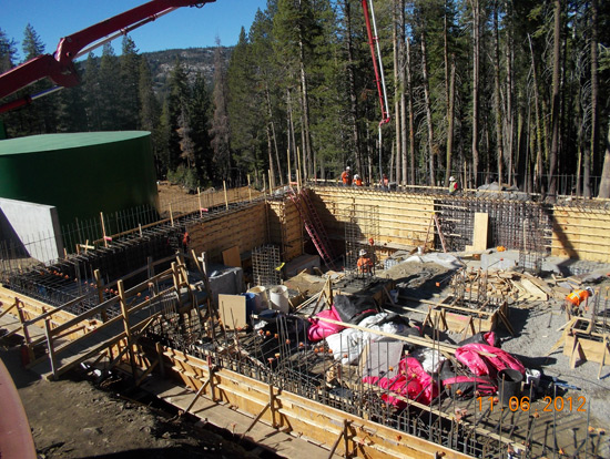 A construction site in a forest with workers, metal frames, and equipment. A green structure is visible in the background.
